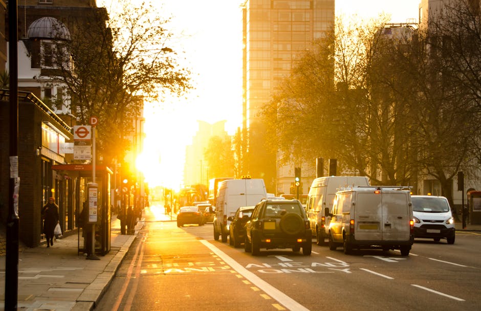 A busy urban street scene during sunset with the sky illuminated in warm orange tones. On the left side, there is a bus stop shelter with a red London Underground sign, and several pedestrians walking along the pavement. To the right, multiple white and dark-colored vans are parked and being loaded or unloaded, indicating a home relocation or furniture transport process. Various cardboard moving boxes and packing materials are visible on the pavement near the vans, some secured with plastic wrap and blankets for protection. The scene is set in a city area lined with trees and tall buildings, including a high-rise structure in the background, with the sunlight casting a golden glow over the scene, emphasizing the busy load and unload activities involved in the moving service provided by Man With a Van Hackney, situated on Kingsland Road.