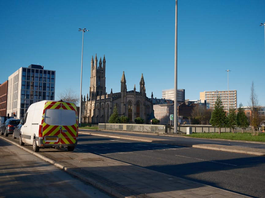 A white panel van with yellow and red reflective chevron markings on the rear doors is parked on the side of a street in Hackney, with a church featuring tall spires and Gothic architecture visible in the background. The van belongs to Man With a Van Hackney, a house removals and furniture transport service. The van is positioned close to a sidewalk with protective pavement edging, and several other vehicles are parked along the street. The surroundings include modern office buildings, residential high-rises, and a row of small, neatly trimmed trees. The sky is clear and blue, indicating daytime. This scene illustrates the logistics involved in home relocation and loading procedures for moving services in the Hackney area, with the van ready to assist with packing, furniture transport, or house removals.