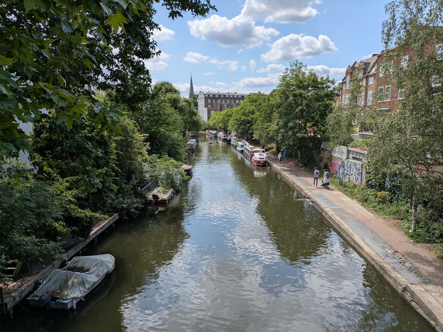 A daytime scene showing a narrow waterway in Hackney lined with green trees on both banks. Several boats are moored along the edges, some covered with protective tarps, while others are open. On the right side, a paved walkway with a few pedestrians runs parallel to the water, with parked cars and a graffiti-covered wall visible further down. In the background, residential buildings with brick facades and large windows are partially obscured by the foliage. The sky is partly cloudy with patches of blue, and the overall setting reflects an urban riverside environment suitable for house removals and furniture transport, as managed by Man With a Van Hackney.