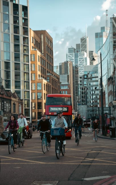 Photograph taken during daytime showing a busy city street in London with modern high-rise buildings and older brick structures. Multiple cyclists, some with backpacks or bags, are riding along the road, with a red double-decker bus displaying route information for Hackney Wick 26 approaching from behind. Pedestrians are walking on the sidewalk, and parked cars can be seen to the sides. The environment is well-lit with natural daylight, highlighting the urban setting. This scene reflects typical transportation and movement in London, relevant to house removals and relocation services offered by Man With a Van Hackney, emphasizing the busy city infrastructure involved in furniture transport and home relocation activities.