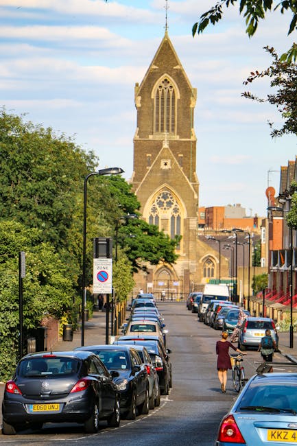 A street scene in Hackney featuring a row of parked cars along the pavement with a woman walking her bicycle and a pedestrian nearby. On the left side, there are trees with green foliage partially covering street lamps and a no-parking sign. In the background, a tall, historic church with Gothic-style windows and a pointed spire dominates the scene, set against a partly cloudy sky. To the right, buildings with retail facades are visible. This image captures the urban environment typical of a home relocation context, highlighting the street’s suitability for furniture transport and moving services, such as those offered by Man With a Van Hackney. The scene illustrates the outdoor loading area where items could be moved from a property to a vehicle, emphasizing the importance of careful planning in the packing and moving process during house removals in the Dalston to London Fields area.