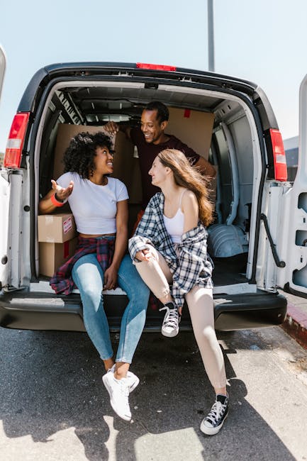 Two young women sit inside the open back of a grey moving van used for house relocation, smiling and engaging in conversation on a bright, clear day. One woman has curly black hair and is wearing a li