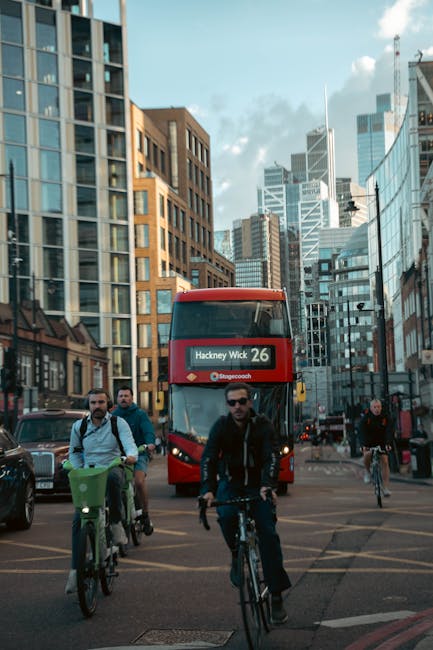 A busy urban street scene in central London during daytime, featuring cyclists riding along the road in front of a red double-decker bus displaying 'Hackney Wick 26' on its electronic destination sign. Several people are cycling, some wearing casual clothing and sunglasses, while others carry backpacks or bags. Modern high-rise office buildings with glass facades and older brick structures line the street, which is filled with traffic and pedestrian activity. The sky is partly cloudy with natural daylight illuminating the scene. This image captures the vibrant city environment typical of a relocation area, highlighting the importance of careful planning and transportation for house removals or moving services. Man With a Van Hackney may utilize such city logistics and urban transportation insights in their professional removals services, ensuring smooth furniture transport and packing for home relocations within this busy setting.