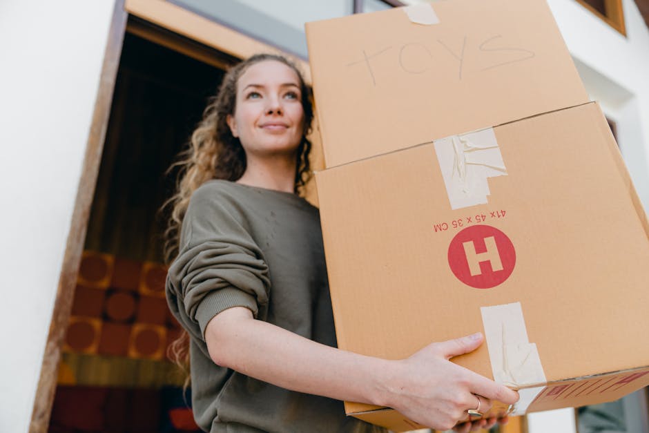 A young woman with curly hair, wearing a grey long-sleeved top, is holding two medium-sized cardboard boxes during a home relocation process inside or just outside a house. The top box has handwritten label 'toys' indicating its contents, while the lower box bears a red and white shipping label with caution symbols and measurements. The woman is positioned near an open doorway, with a wooden frame visible, and is smiling slightly as she prepares for furniture transport or packing and moving activities. The background includes interior elements with soft lighting, and the scene reflects the logistics of house removals with the woman actively involved in loading or transporting items, supported by professional moving services like Man With a Van Hackney.