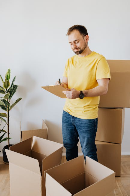 A young woman with shoulder-length blonde hair, wearing a casual green T-shirt, ripped jeans, and black sneakers with pink laces, stands inside a room next to a stack of three large cardboard moving boxes; the top box is open with flaps folded back, and she is holding a pen and a small notepad, appearing to take notes or check an inventory. Behind her, there is a potted plant with broad green leaves placed in a woven basket, and the floor is wooden with a light finish. The scene suggests she is involved in packing or organising items for a home relocation, with natural lighting illuminating the space, which is part of the house removal process supported by Man With a Van Hackney.