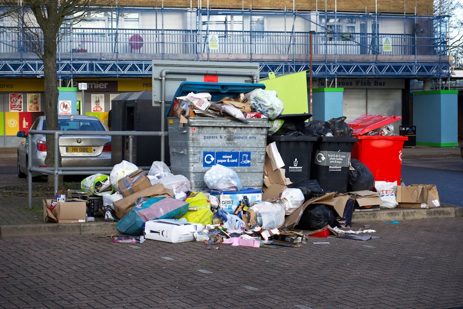 A large pile of mixed household waste and recycling, including cardboard boxes, plastic bags, black refuse sacks, and various paper and packaging materials, is overflowing from multiple recycling bins