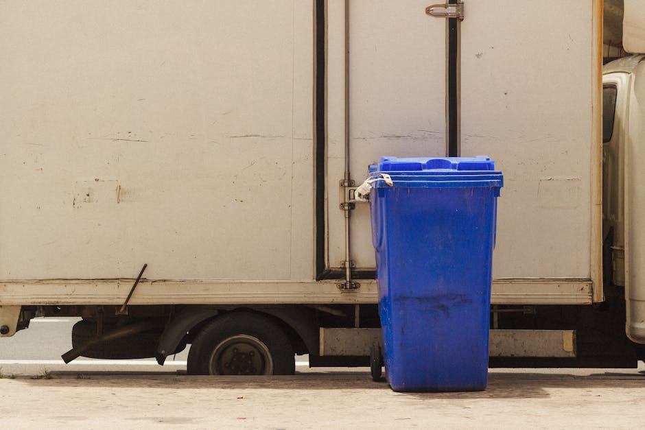 A large pile of mixed household waste and recycling, including cardboard boxes, plastic bags, black refuse sacks, and various paper and packaging materials, is overflowing from multiple recycling bins